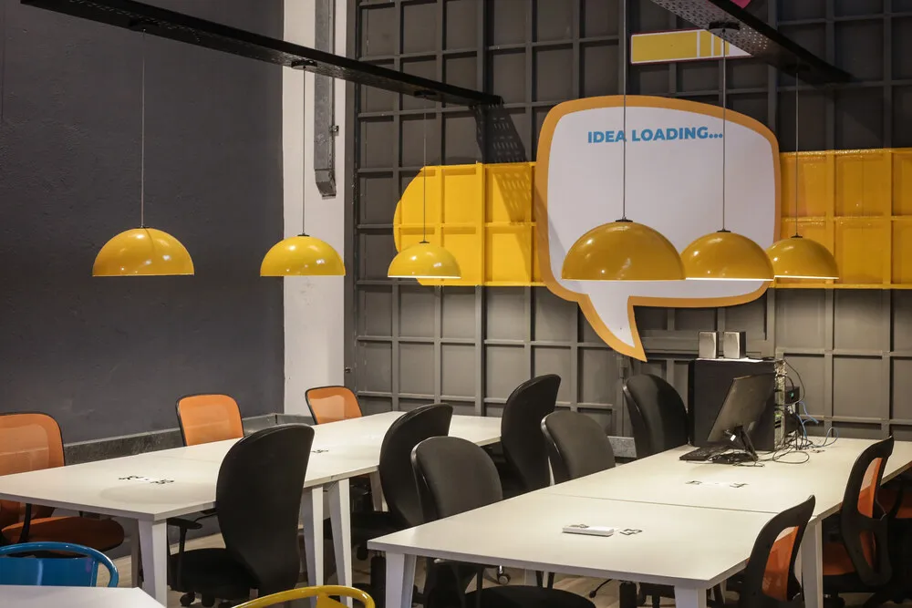 Sala de trabajo con escritorios blancos, sillas naranjas y lámparas colgantes amarillas en Host Orfeo