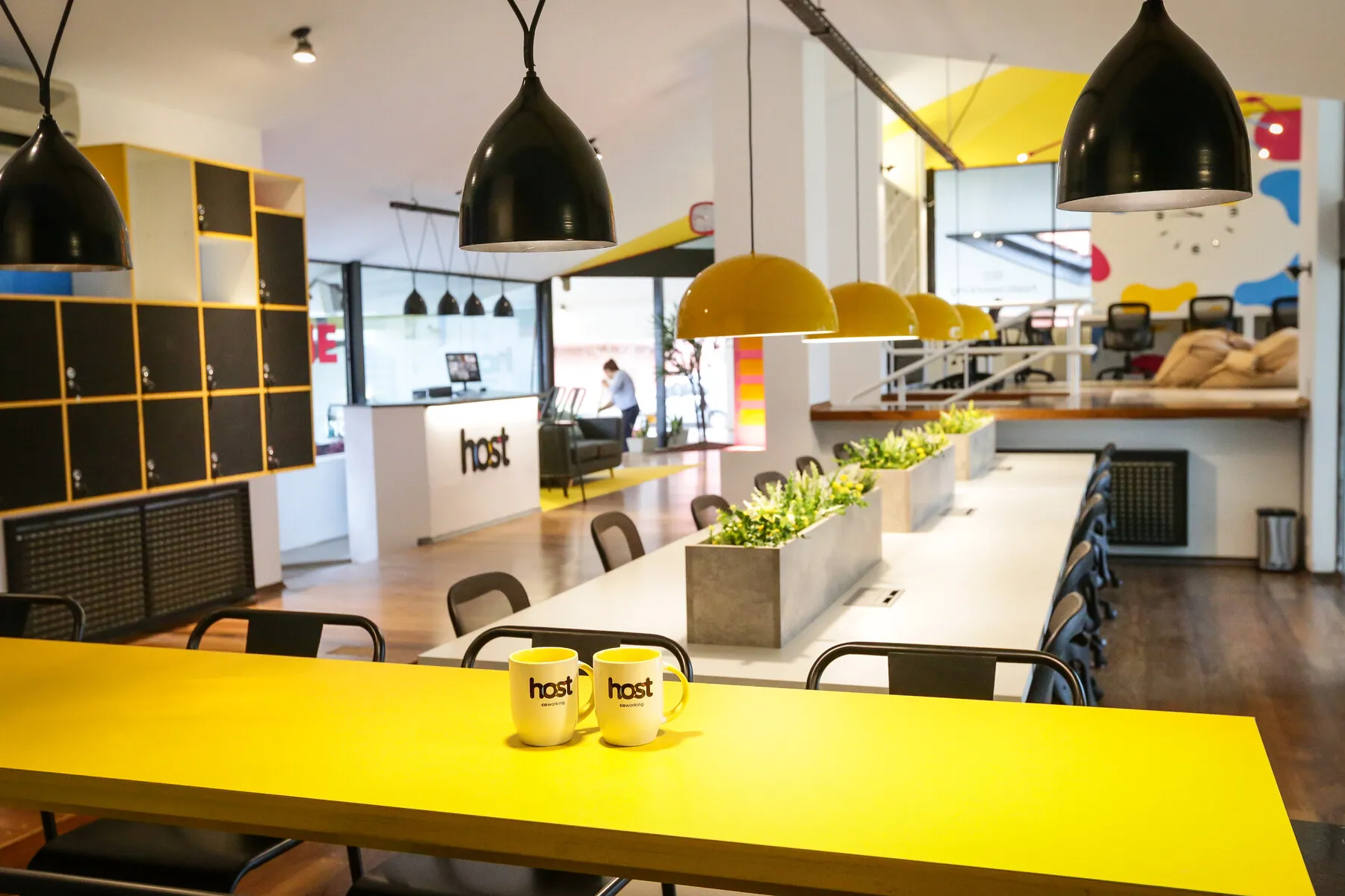 Recepción de Host Coworking en Villa Belgrano, con mesa amarilla, tazas de Host, lockers y mostrador de recepción