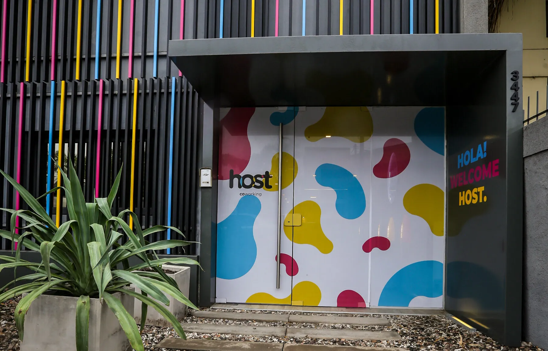 Fachada de Host Comunidad de Negocios en Orfeo, con puertas de vidrio de colores y el cartel HOLA WELCOME HOST